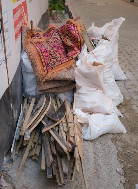 A collection of discarded household items and construction debris piled along a paved sidewalk beside a building wall. The pile includes a patterned fabric rug with vibrant pink, purple, and orange to