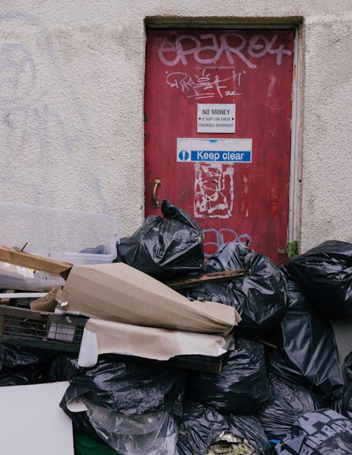 A collection of discarded household items and construction debris piled along a paved sidewalk beside a building wall. The pile includes a patterned fabric rug with vibrant pink, purple, and orange to