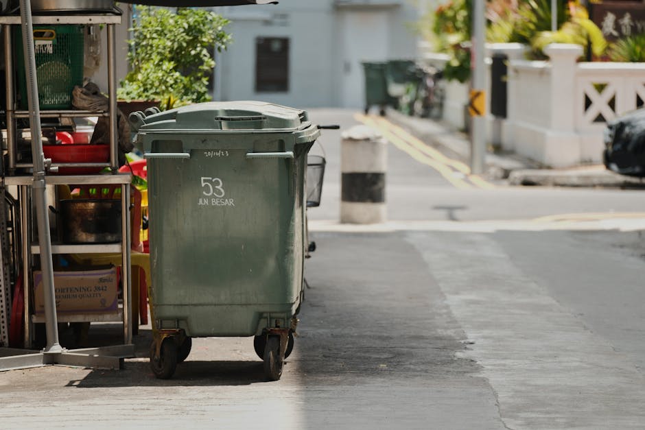 A black metal utility cart with large rear wheels and a flat platform in an urban alleyway, filled with various types of waste including plastic bottles, bags, and other debris. The cart is positioned